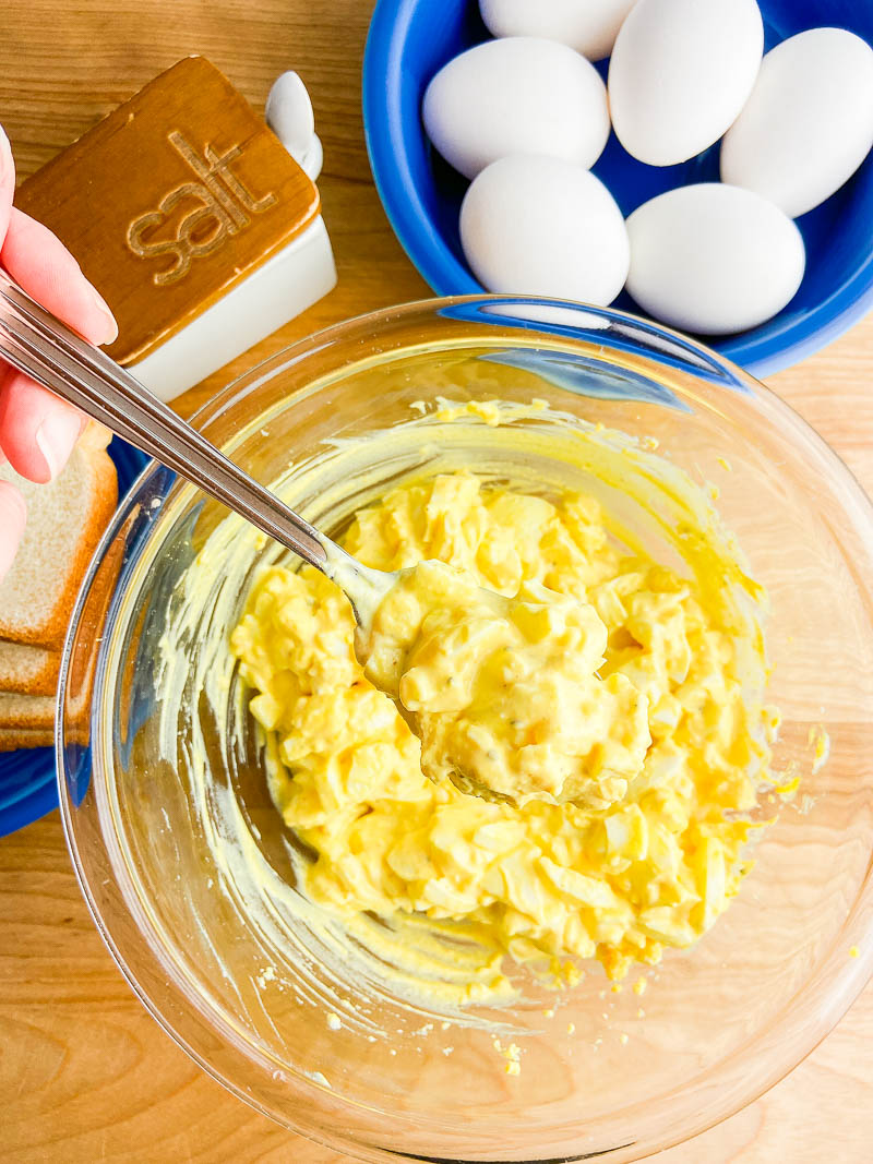 A spoonful of egg salad over a glass bowl on a wooden cutting board with a bowl of eggs. 