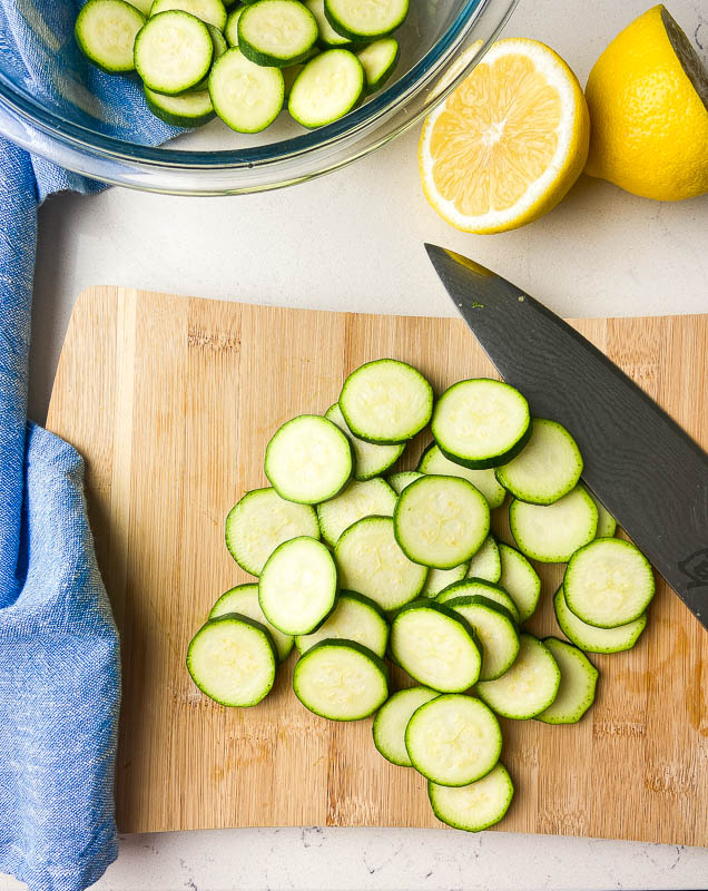 Sliced zucchini on a wooden cutting board with a lemon and blue napkin. 