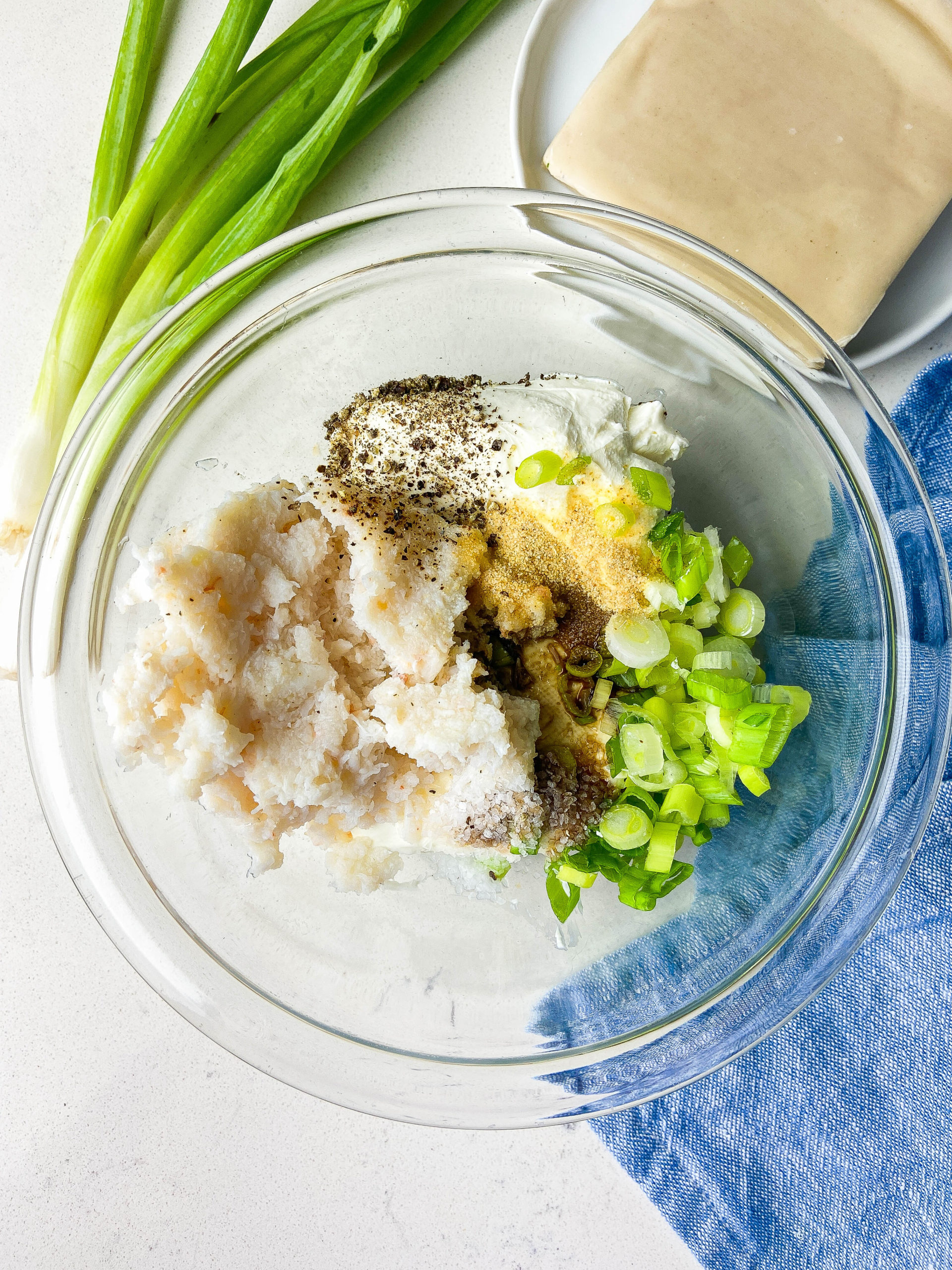 Overhead photo of ingredients in crab wontons in a glass bowl. Green onions, wonton wrappers and a blue napkin in the background. 