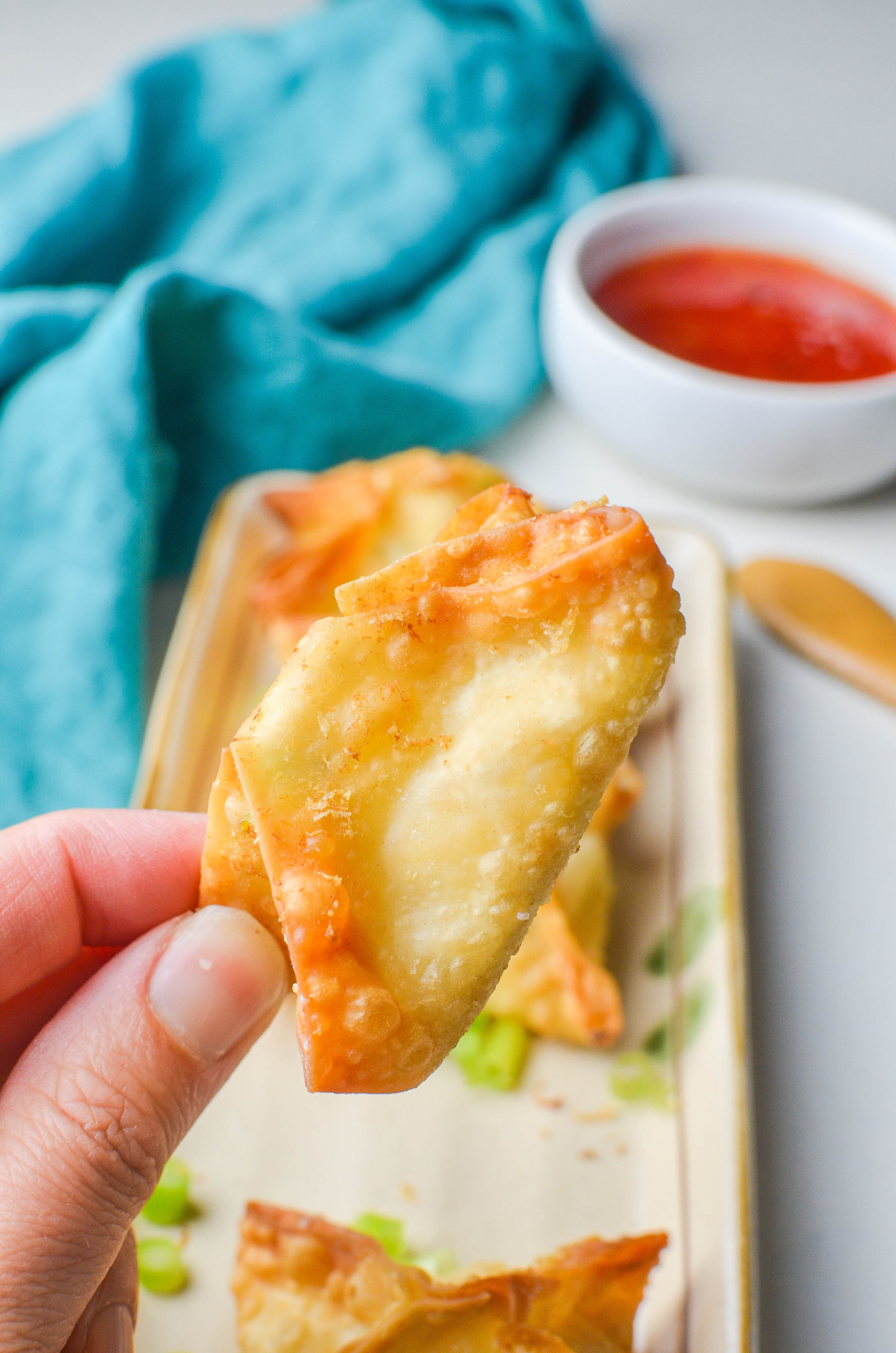 Hand holding a crab wonton with teal napkin and sweet thai chili sauce in the background. 