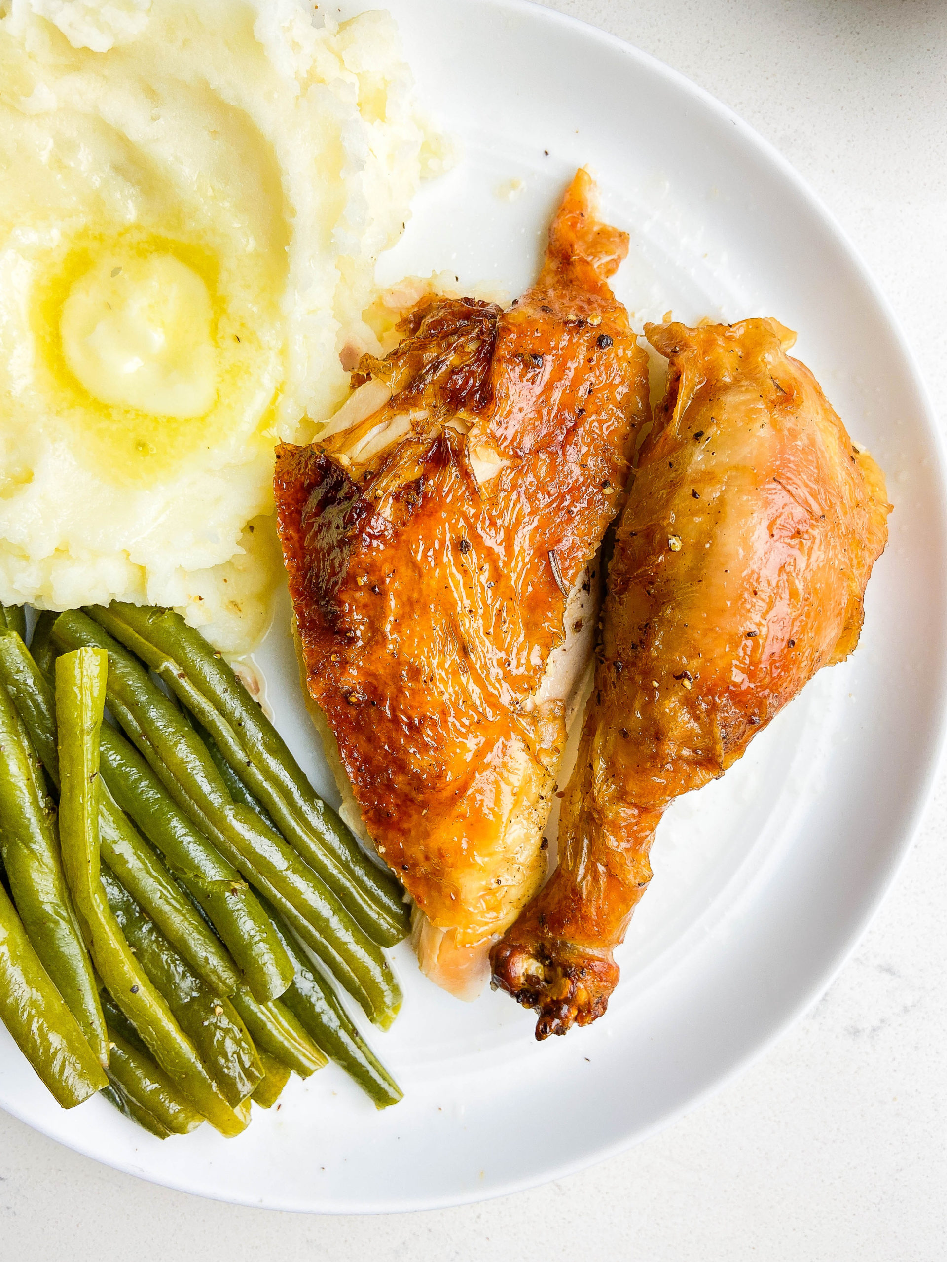 Roast chicken on a white plate with green beans and mashed potatoes. 