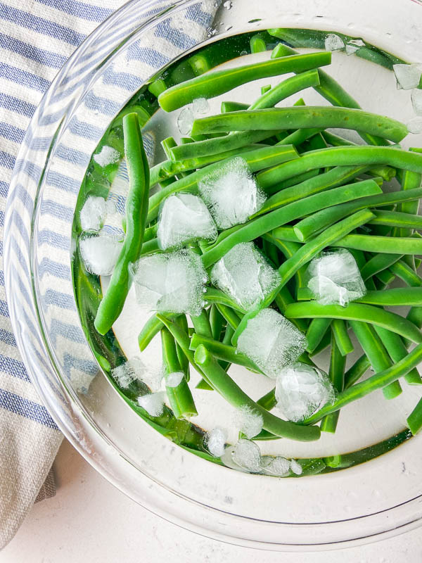 Green beans in an ice bath in a clear bowl with a blue striped towel. 