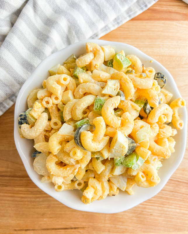 Overhead photo of macaroni salad in a white bowl on a wooden cutting board. A stripped towel off to the side. 