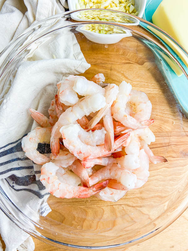 Raw shrimp in a clear bowl on a wooden cutting board. 