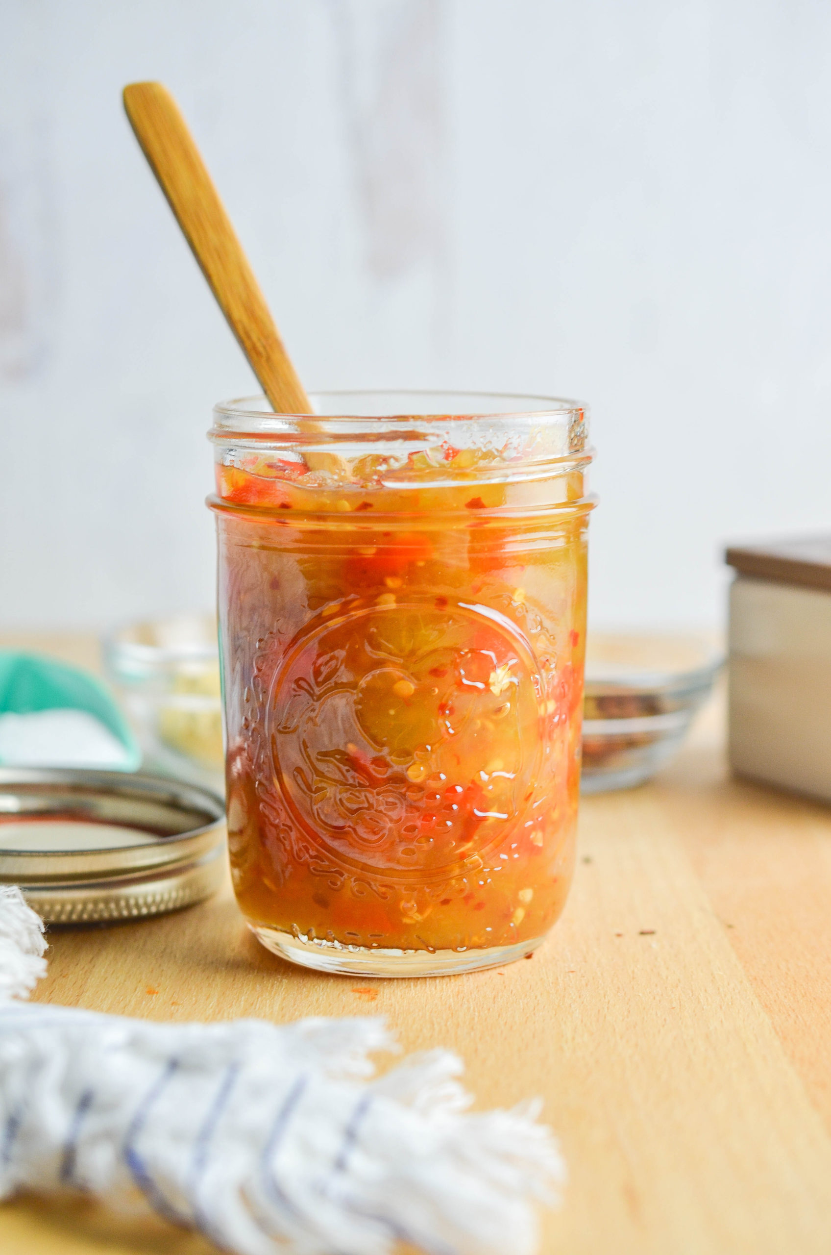 Tomato relish in a glass jar with a wooden spoon on a wooden cutting board with white background. 