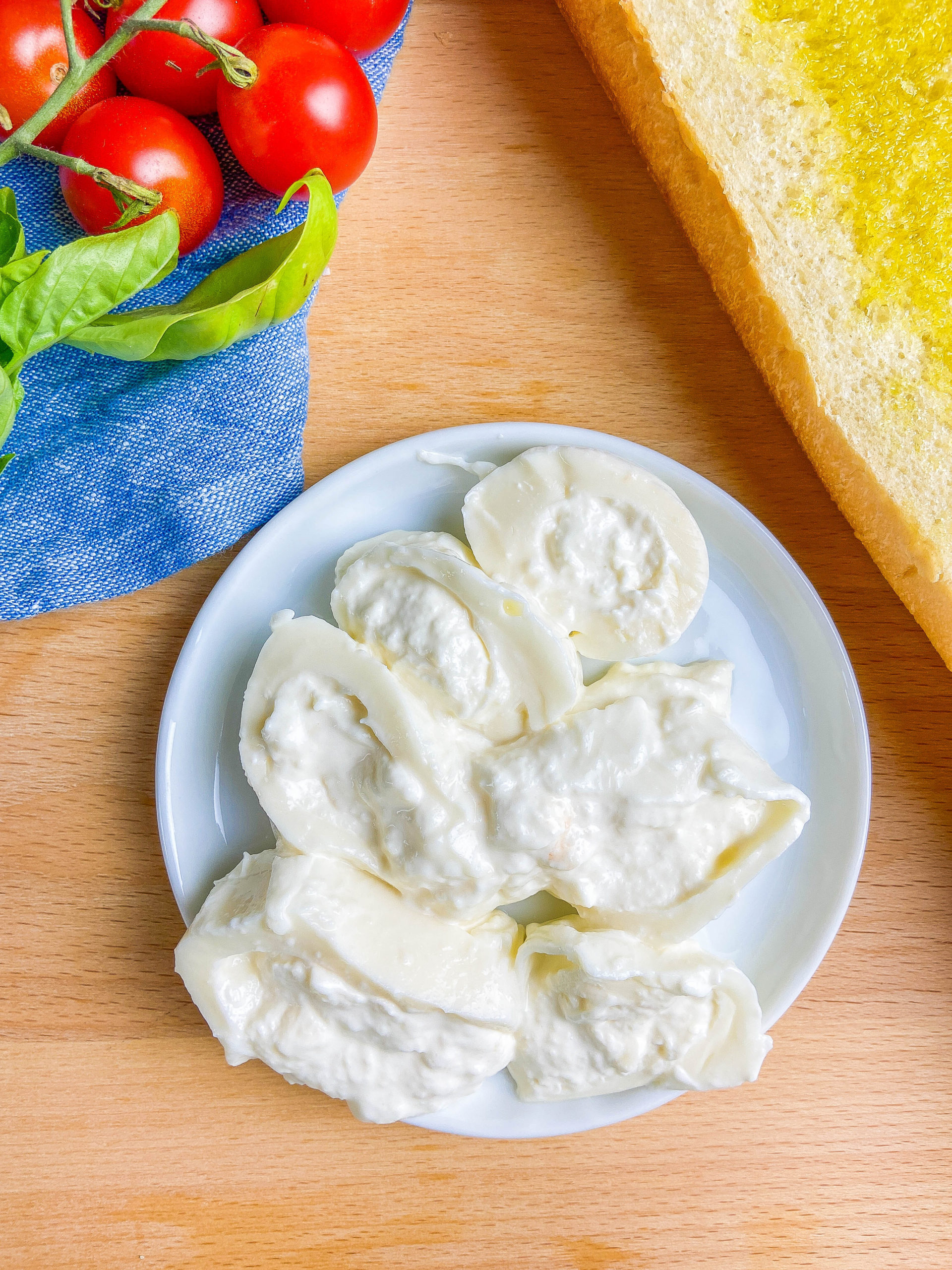 Overhead photo of burrata cheese on a white plate. Wooden cutting board background with blue towel, bread, cherry tomatoes and basil. 