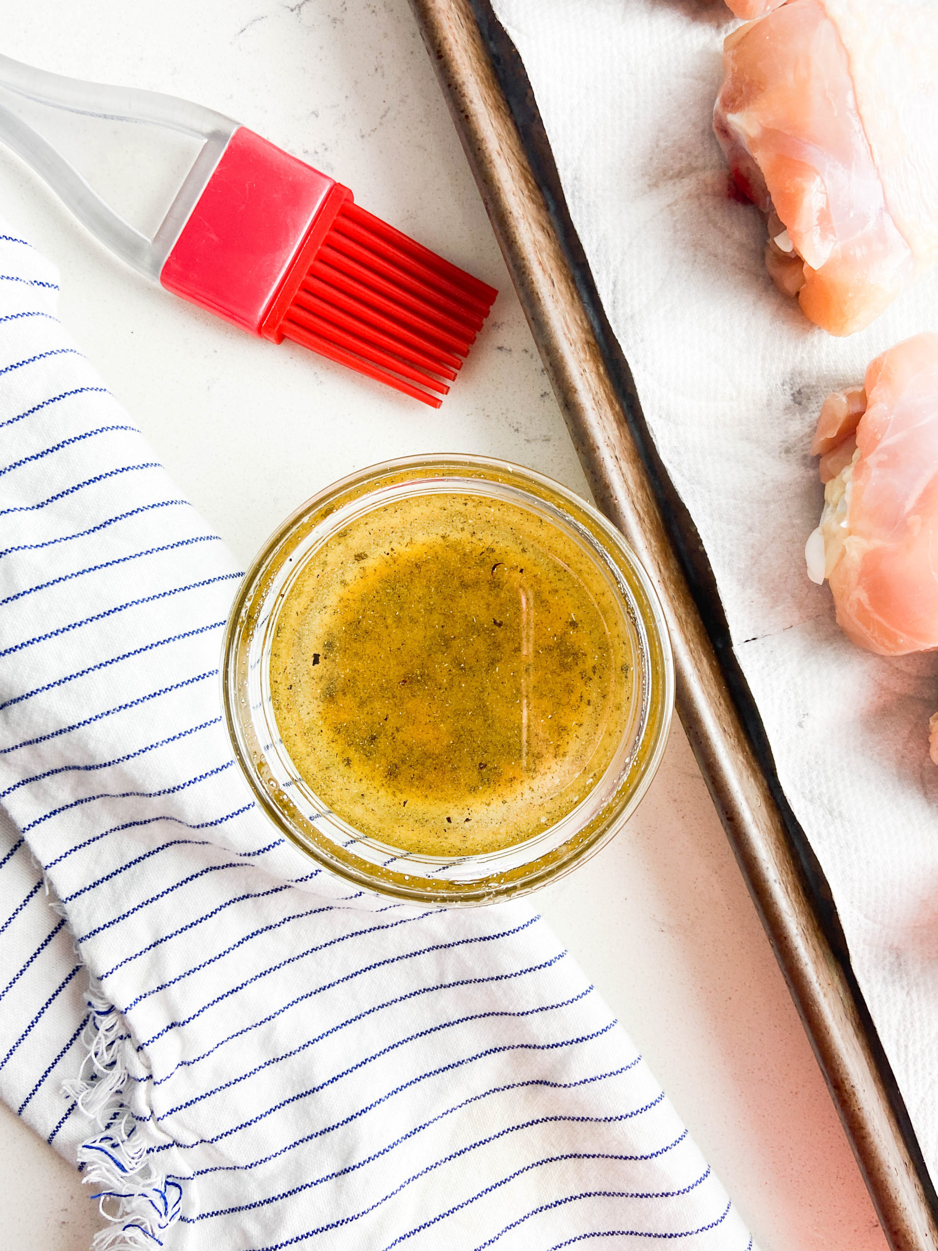 Garlic pepper oil in a clear glass bowl next to a striped towel on a white background. Chicken and pastry brush off to the side. 