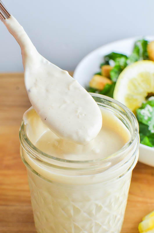 A spoonful of Caesar dressing over a mason jar with a caesar salad in the back ground. 