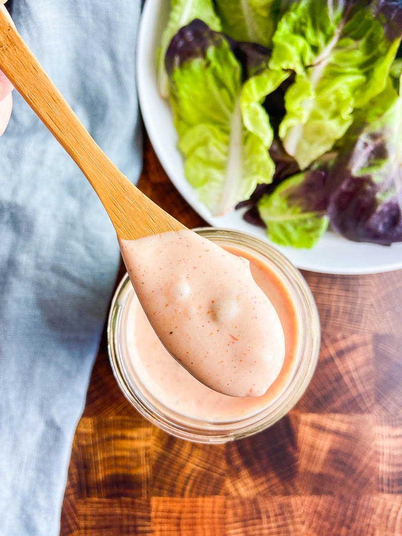 Overhead photo of thousand island dressing on a wooden spoon with a wooden background and salad. 