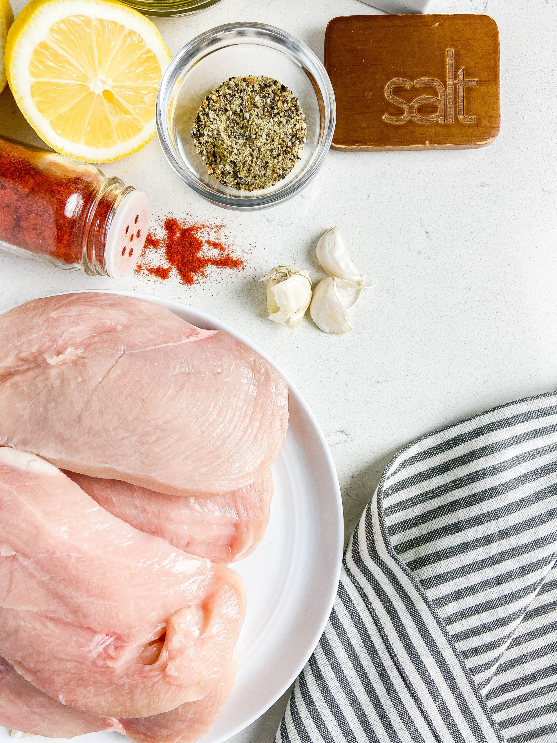 Raw chicken breasts on a plate with paprika, garlic, lemon pepper, lemon and a striped napkin. 