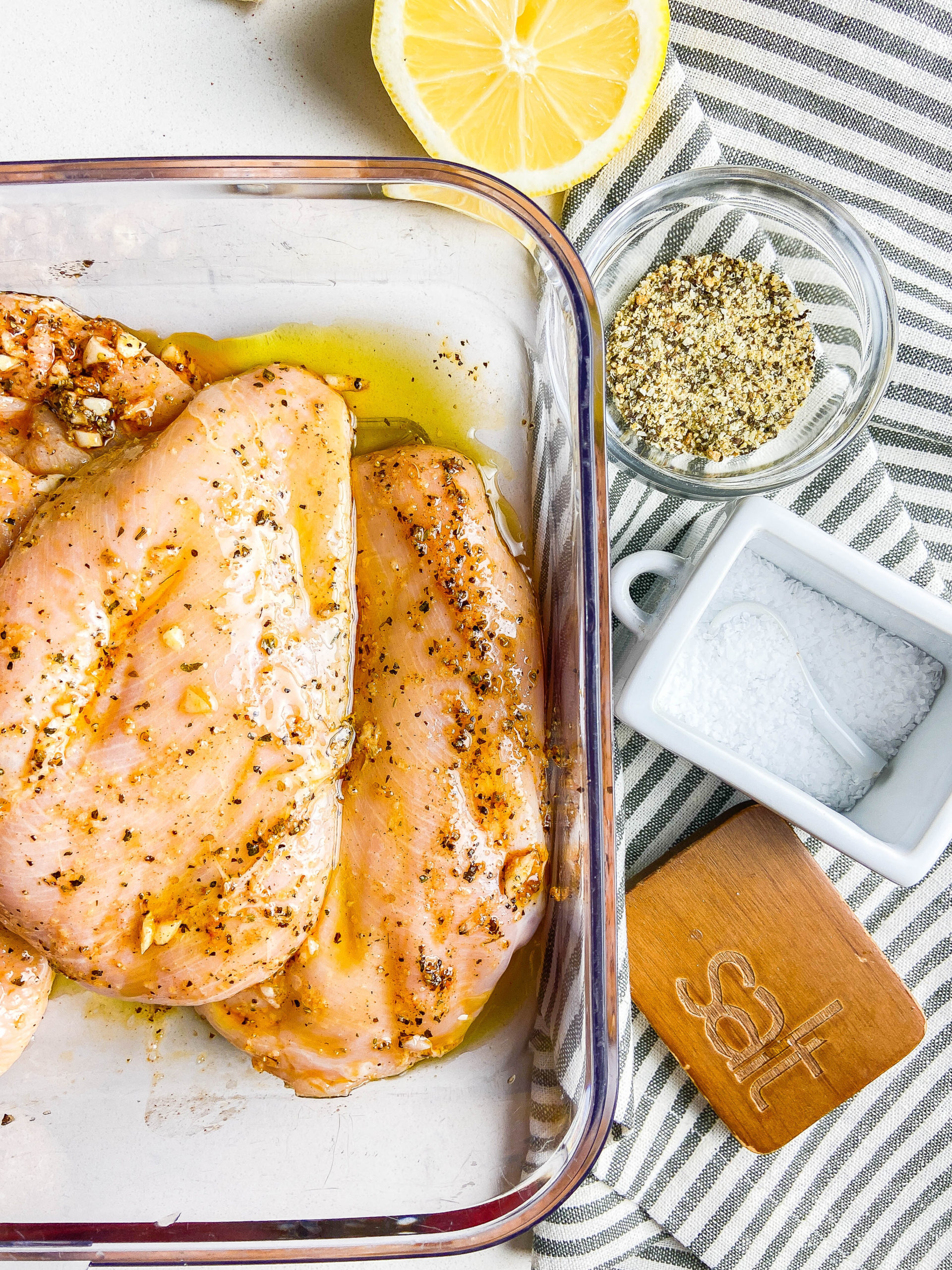 Overhead photo of chicken marinating in lemon pepper seasoning in olive oil. Stripped towel with salt, pepper and lemon pepper seasoning,