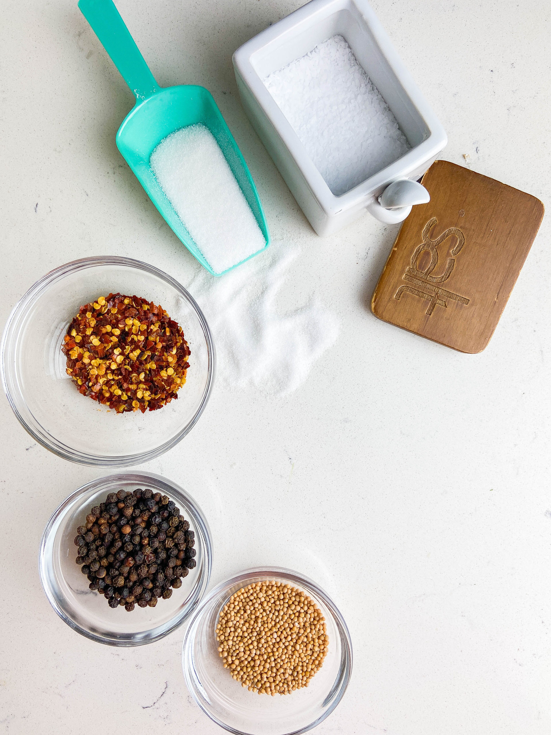 Mustard seeds, black peppercorns, crushed red peppers in glass bowls. Sugar in a teal scooper and salt in a white salt cellar. 