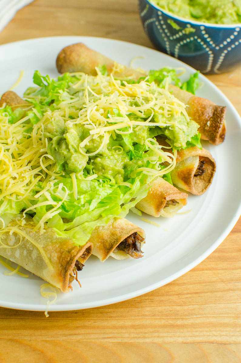Shredded beef taquitos topped with lettuce, cheese and guacamole on a white plate. 