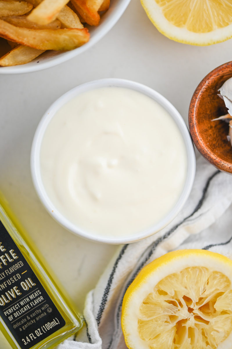 Overhead photo of truffle aioli in a white bowl with lemons, oil and fries.